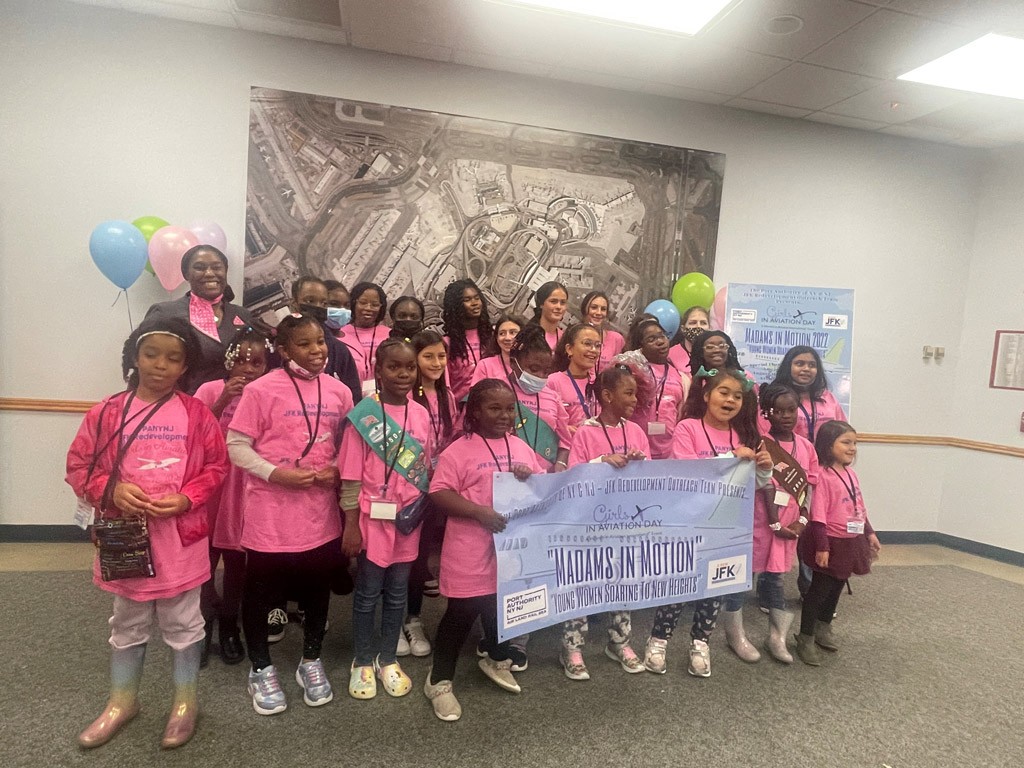 Group of young girls in pink shirts holding a banner for 'Madams in Motion' event at JFK.