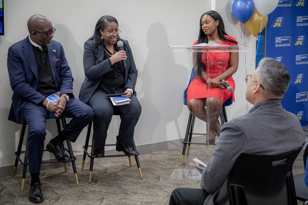 A panel discussion featuring three speakers, one of whom is at a podium. The backdrop includes a blue and gold design.