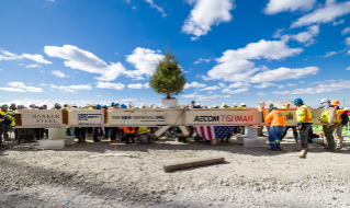 Construction workers gathered around a steel beam with logos, commemorating the new terminal at JFK International Airport, highlighted by a small tree.