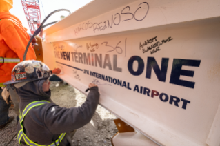 A construction worker wearing a hard hat signs a beam labeled 'THE NEW TERMINAL ONE JFK INTERNATIONAL AIRPORT,' surrounded by various signatures.