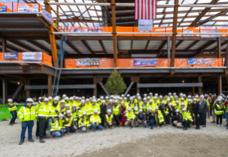 A large group of construction workers and associates, wearing safety helmets and yellow reflective vests, pose together at a construction site with an American flag above them.