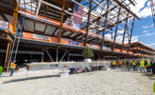 Construction site with workers gathered around a beam decorated with signatures and a tree, under a partially constructed metal frame, an American flag visible overhead.