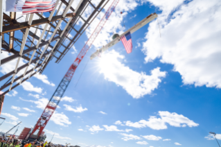 A large crane lifts a steel beam adorned with an American flag against a bright blue sky filled with clouds. Construction workers gather below to observe the event.