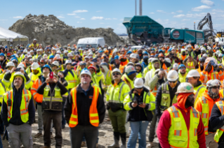 A large crowd of construction workers in bright safety vests and hard hats gathers outdoors, capturing photos of an event.