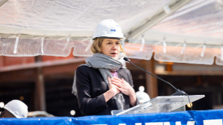 A woman wearing a hard hat and scarf speaks at a podium during a construction event.