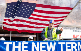 A man in a hard hat and high-visibility vest speaks passionately at a podium, with an American flag in the background and a banner reading 'THE NEW TERM.'