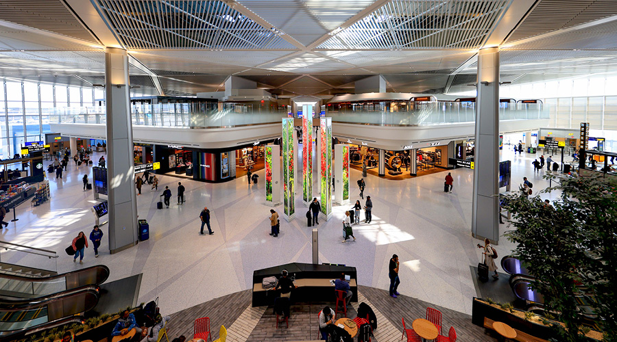 A spacious airport terminal with passengers walking around, featuring large glass windows and colorful installations.