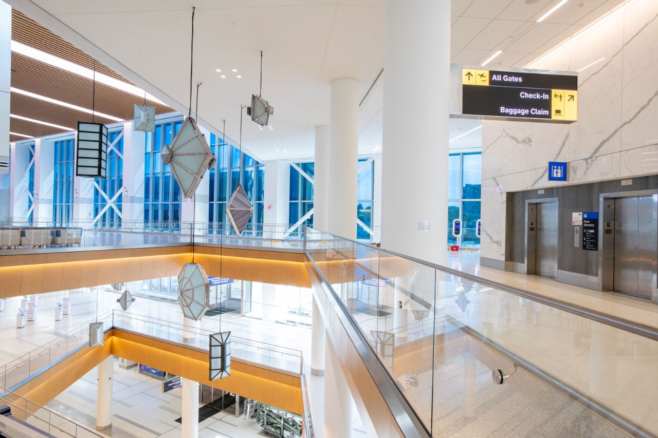A modern airport terminal interior featuring suspended geometric light fixtures, glass railings, and clear signage for gates and services.