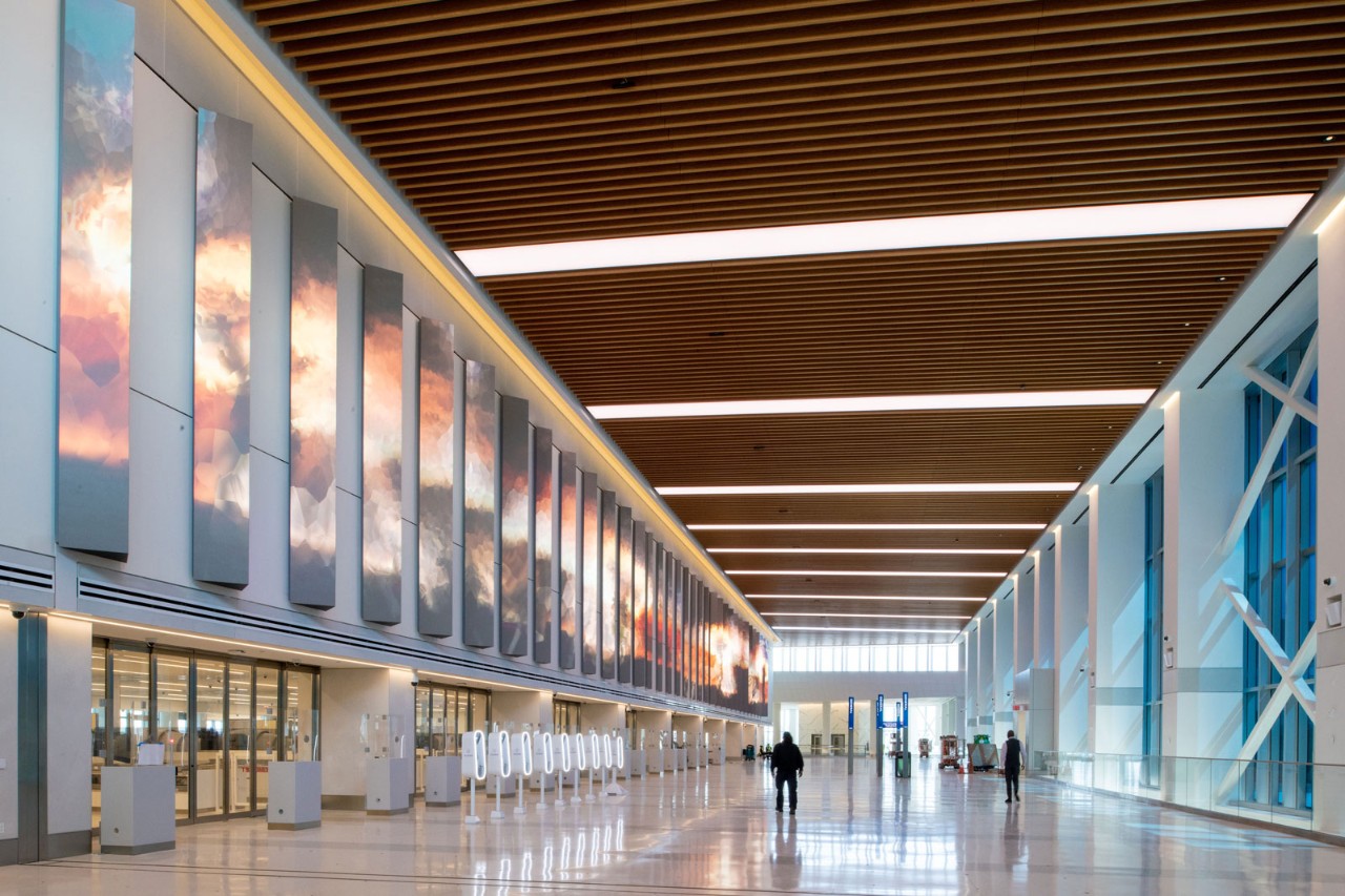 A modern airport interior featuring a long corridor with large digital displays and wooden beams overhead. The space is well-lit and mostly empty, with a few people walking in the distance.