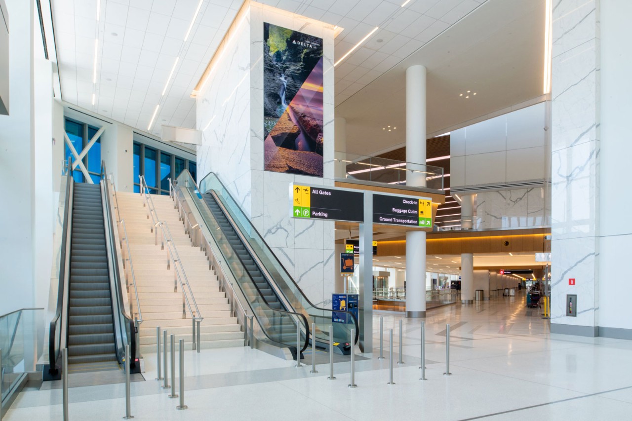 A modern airport interior featuring escalators, a large digital display, and directional signage.