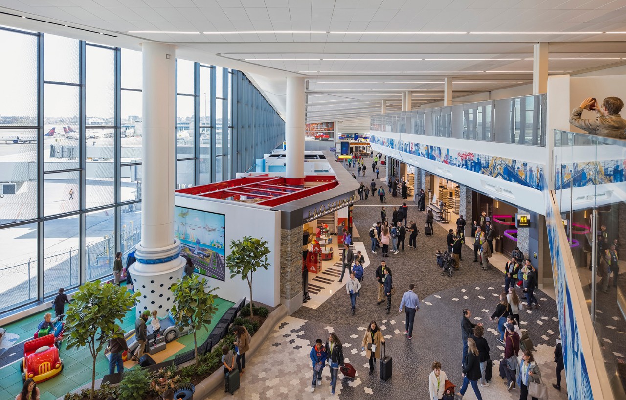 A spacious airport terminal filled with travelers, featuring large windows and various shops, with play areas for children visible.